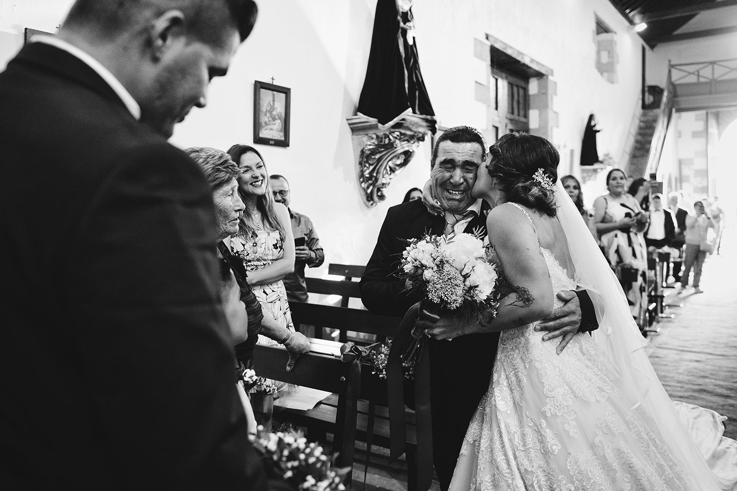 Novia besando emocionada a su padrino durante una ceremonia religiosa de boda en Fuerteventura, rodeada de familiares e invitados.