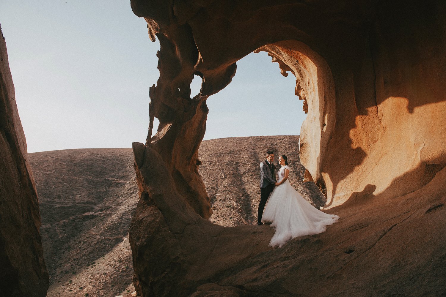 Novios cogidos de la mano durante su sesión de postboda en el Arco de Las Peñitas, Fuerteventura, enmarcados por las formaciones rocosas.